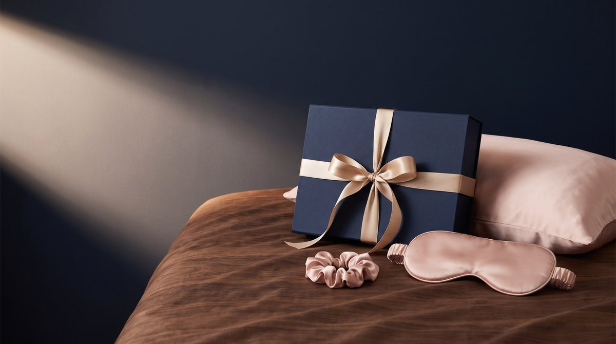A silk pillowcase, a silk sleep mask and a silk scrunchie folded inside an open gift box on a linen bedspread