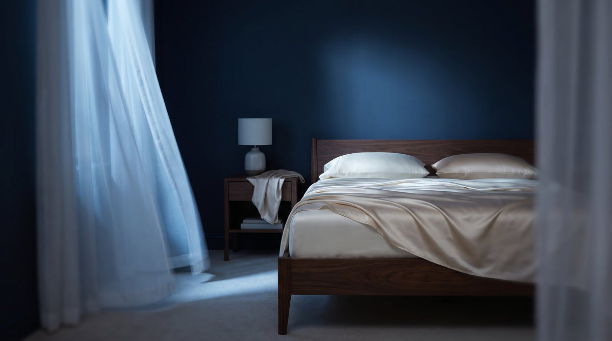 A pale grey silk flat sheet turned down on a bed beside an open window with a light curtain moving in the breeze