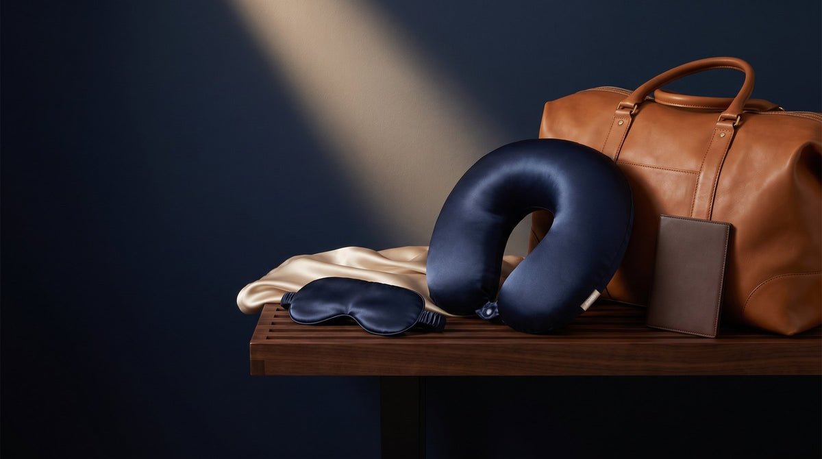 A silk sleep mask and a silk covered neck pillow beside a small travel pouch on a linen surface
