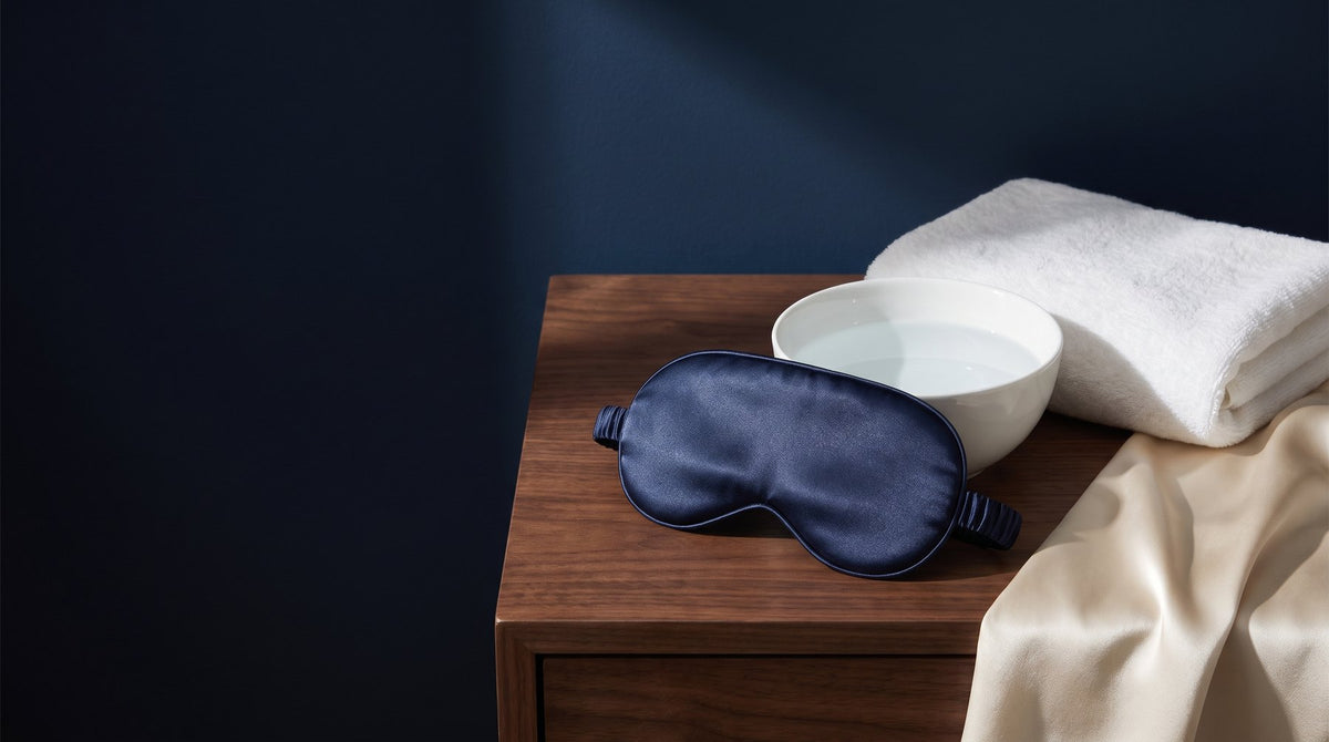A silk sleep mask lying flat on a folded white towel next to a small basin of water