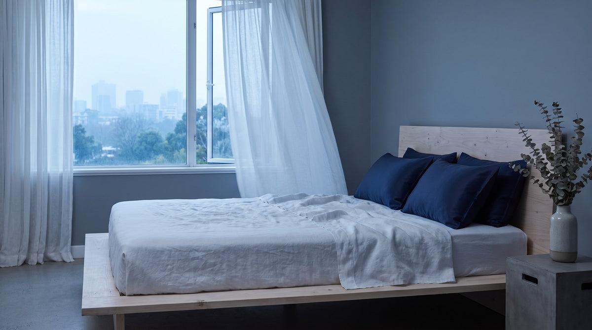 A bed with navy silk pillowcases next to an open window with sheer curtains in cool morning light
