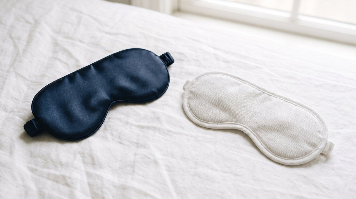 A navy silk sleep mask next to a white cotton sleep mask on a linen sheet