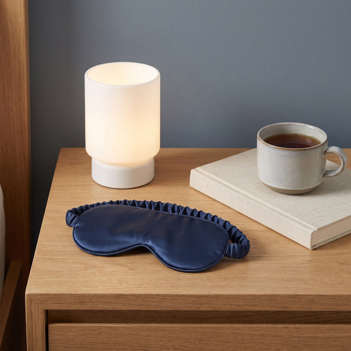 Navy KINT classic silk sleep mask on a bedside table with a lamp, a book and a cup of tea