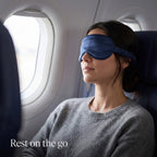 Woman resting in an airplane window seat wearing the navy KINT classic silk sleep mask